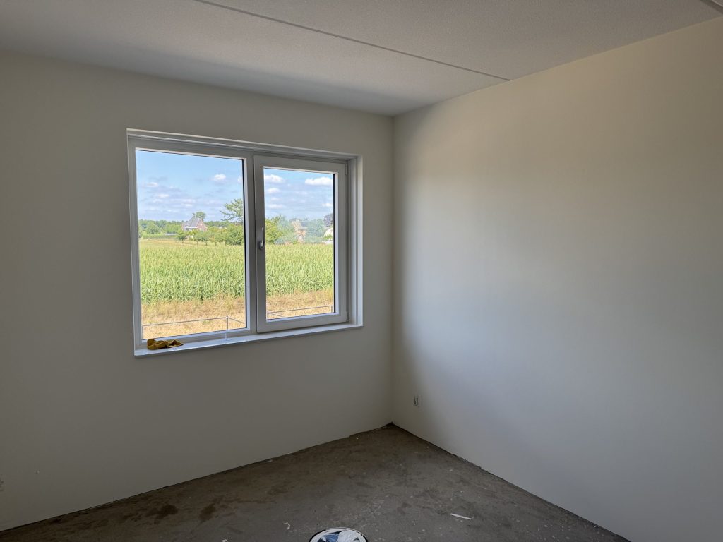 Lege slaapkamer in een nieuwbouwhuis met gladde, lichte wanden en uitzicht op een maisveld